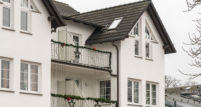 Three-story residential building with white exterior, featuring a sloped roof, multiple windows, and a balcony adorned with festive decorations.