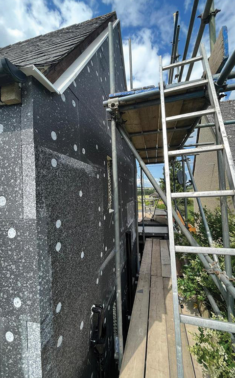 Scaffolding set up next to a house undergoing exterior renovation, featuring a textured black wall with circular white patches, against a blue sky with clouds.