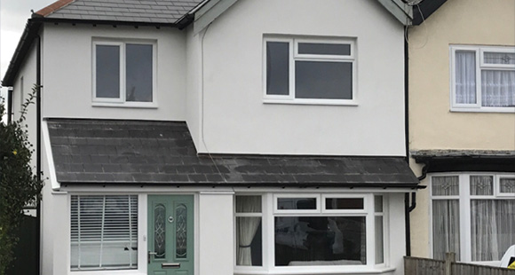 A modern two-story house with a white exterior and a grey slate roof, featuring large windows and a green front door, surrounded by a neighboring house. Ideal for family living and home buyers.