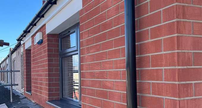 Close-up of a brick wall featuring a window, with a black downpipe and scaffolding in the foreground, showcasing the details of modern construction and red bricks.